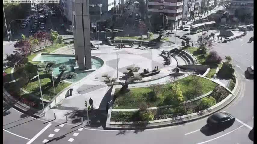 A plaza with a circular design featuring landscaping, water features, and benches, surrounded by roads and buildings on a sunny day.