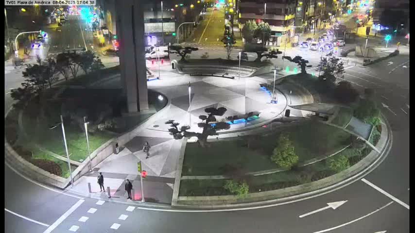 A nighttime aerial view of a roundabout with a geometrically patterned plaza in the center, surrounded by landscaping and streetlights, with a few pedestrians and cars visible.