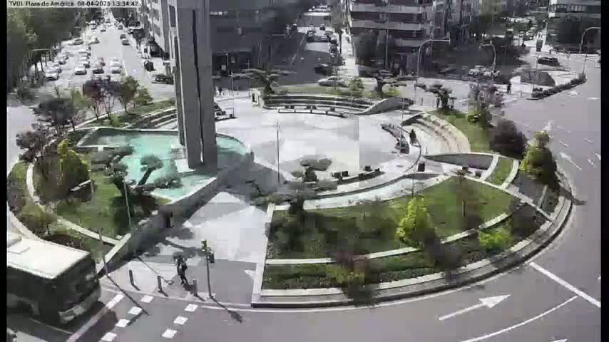A circular plaza with a fountain and landscaping is surrounded by streets and buildings on a sunny day.
