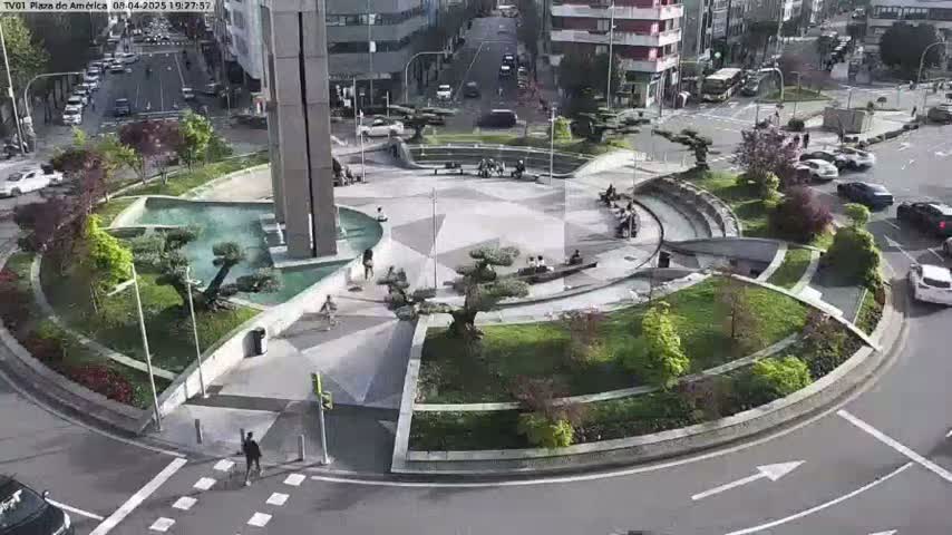 A paved plaza with a central fountain and landscaping is surrounded by a traffic circle on a sunny day.