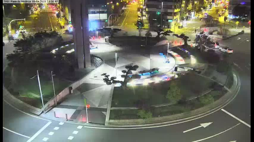 A nighttime aerial view of a plaza with geometric landscaping, streetlights, and sparse vehicle traffic on surrounding roads.