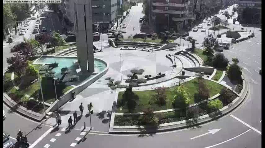 A sunny day reveals a circular plaza with landscaping, a fountain, and a few pedestrians, situated at a roadway intersection.