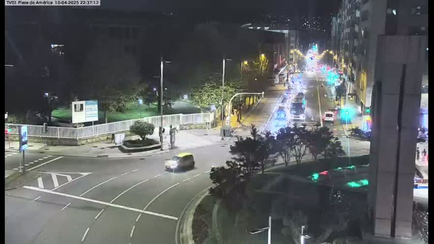 A nighttime view of a street intersection with cars driving on a multi-lane road surrounded by buildings and trees.