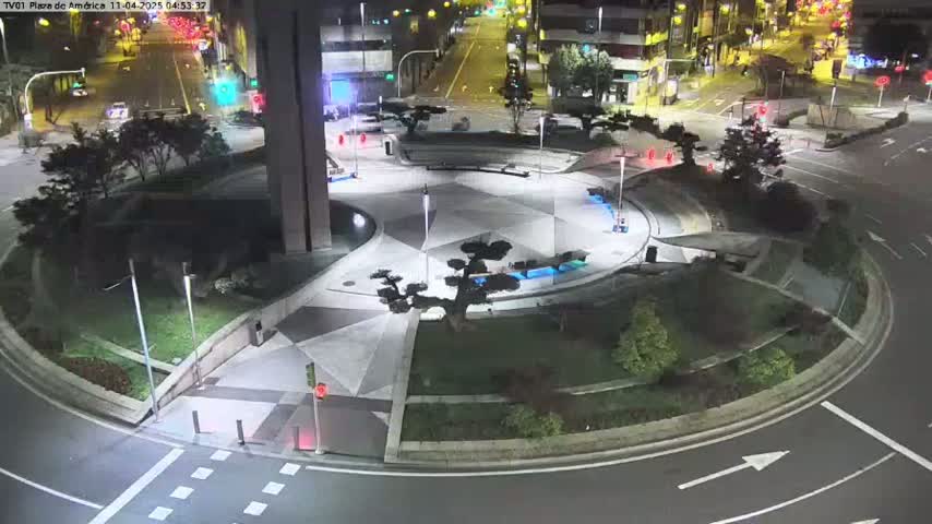 A nighttime aerial view of a circular plaza with geometric landscaping, a small tree in the center, and surrounding roads with minimal traffic.