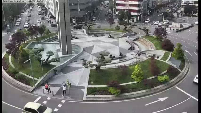 A circular plaza with geometric landscaping, a central fountain, and several small trees is surrounded by a roadway with cars, under an overcast sky.