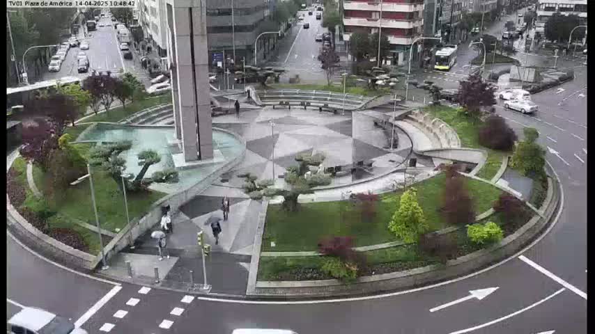 A circular plaza with landscaped gardens and a water feature is situated in the middle of a roundabout on a rainy day.