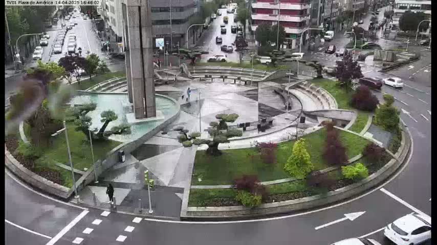 A wet roundabout features a modern fountain and landscaping, surrounded by city streets with cars and buildings.