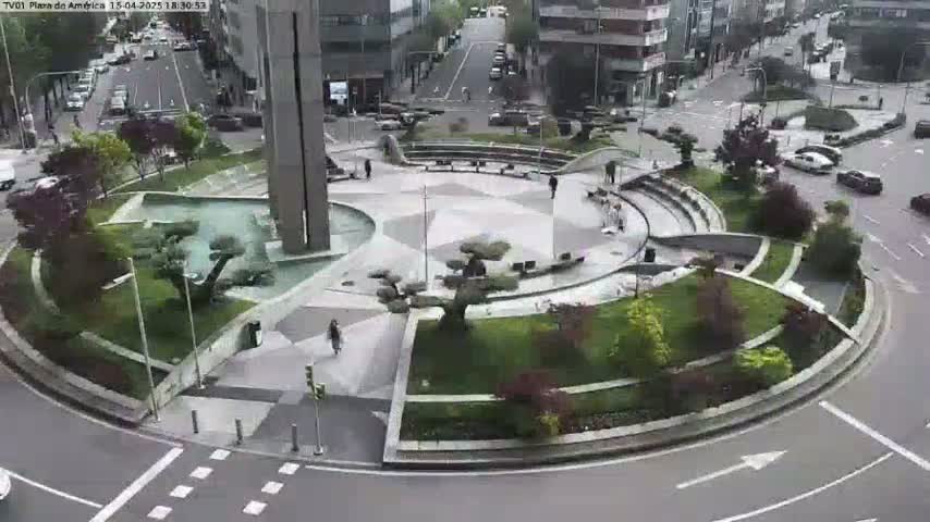 A circular plaza with landscaping, a fountain, and a tall building is visible, with cars driving on roads surrounding it under a partly cloudy sky.