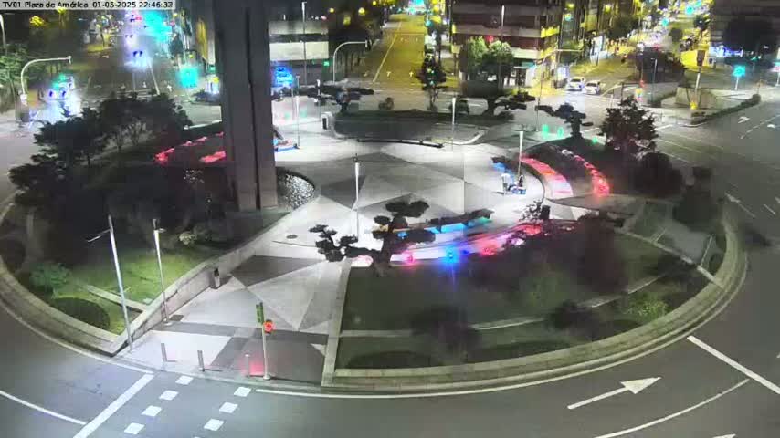 A nighttime aerial view of a roundabout with landscaped areas, streetlights, and a few vehicles on surrounding roads.