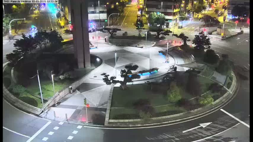 A nighttime aerial view shows a circular plaza with geometric landscaping, small trees, and blue-lit features, surrounded by roadways and buildings.