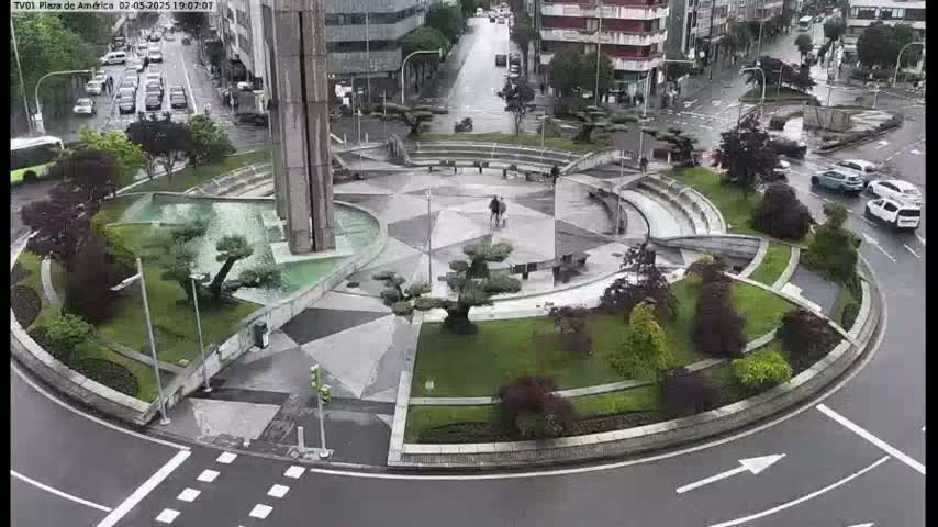 A rain-slicked roundabout features a central plaza with landscaping, a fountain, and a modern monument, surrounded by city streets with cars.