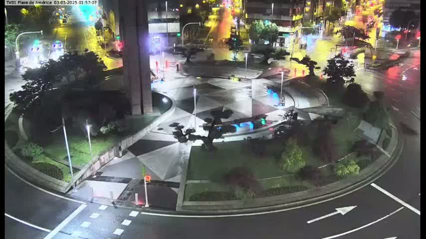 A nighttime, wet plaza with geometric landscaping and several trees is surrounded by a circular roadway with some vehicle traffic.