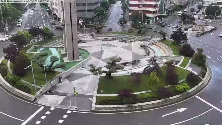 A circular plaza with landscaped gardens and benches is surrounded by a roadway on a rainy day.
