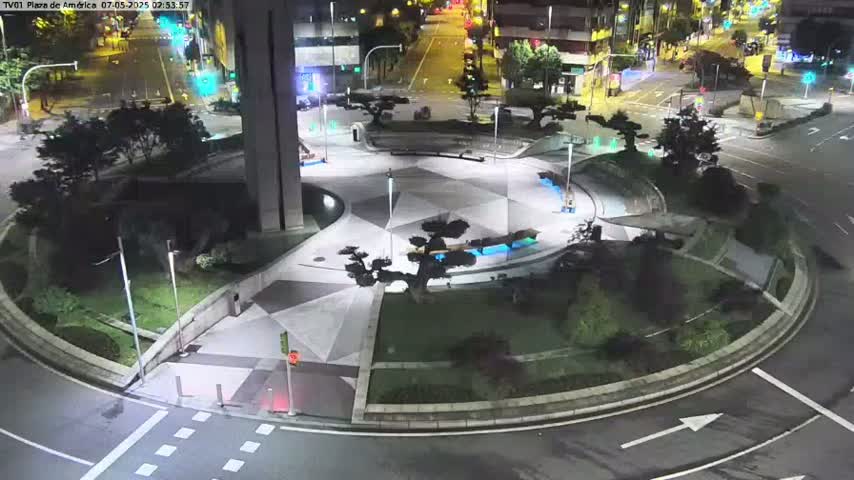 A nighttime aerial view of a circular plaza with geometric paving, landscaping, and streetlights, surrounded by roads and buildings.