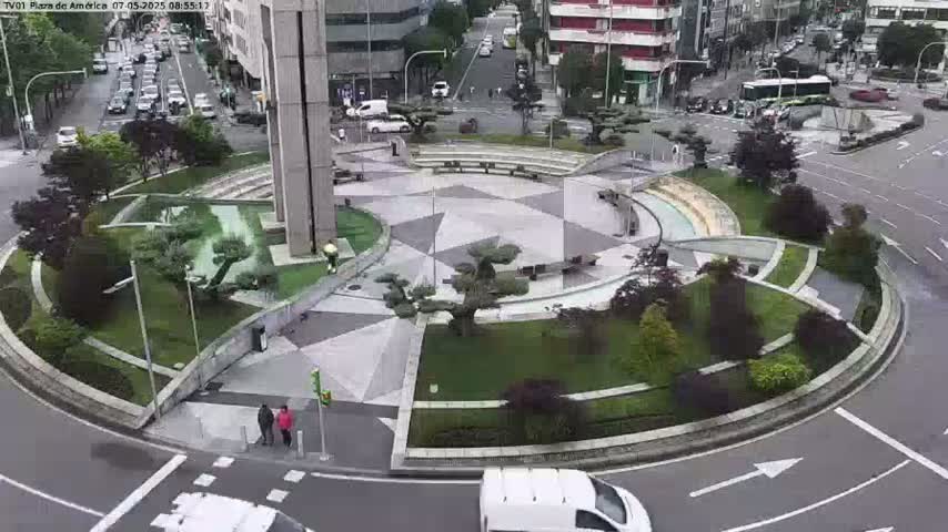 A circular plaza with landscaped gardens and a geometric pattern in the center is surrounded by a roadway with cars and buildings in the background, under overcast skies.