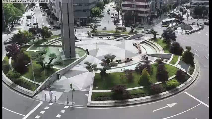 A landscaped traffic circle with several trees and shrubs is situated in a city, with cars visible on surrounding roads under a mostly clear sky.