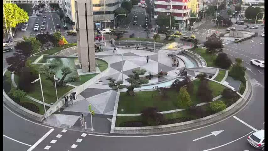 A paved plaza with geometric landscaping, a central fountain, and surrounding trees is situated in a roundabout with cars driving around it at night.