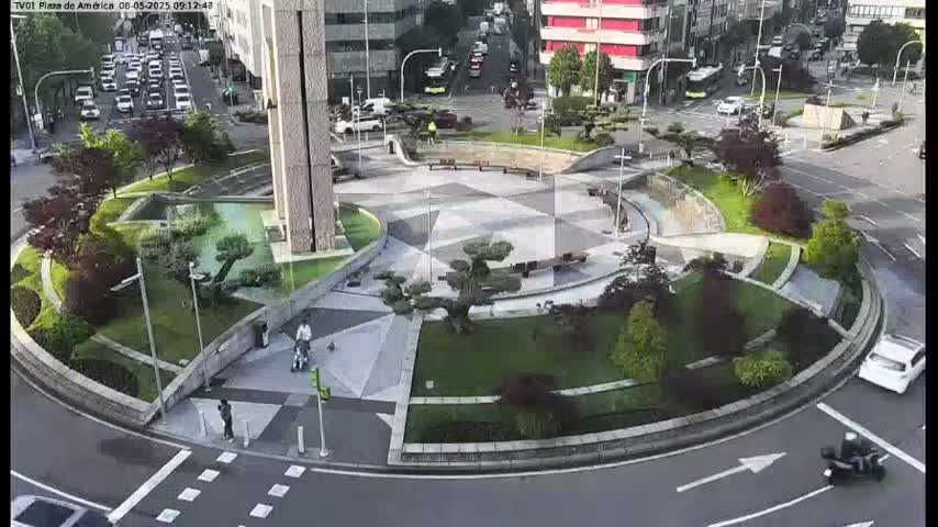 An aerial view shows a roundabout with landscaped gardens and a few pedestrians, under a mostly sunny sky.