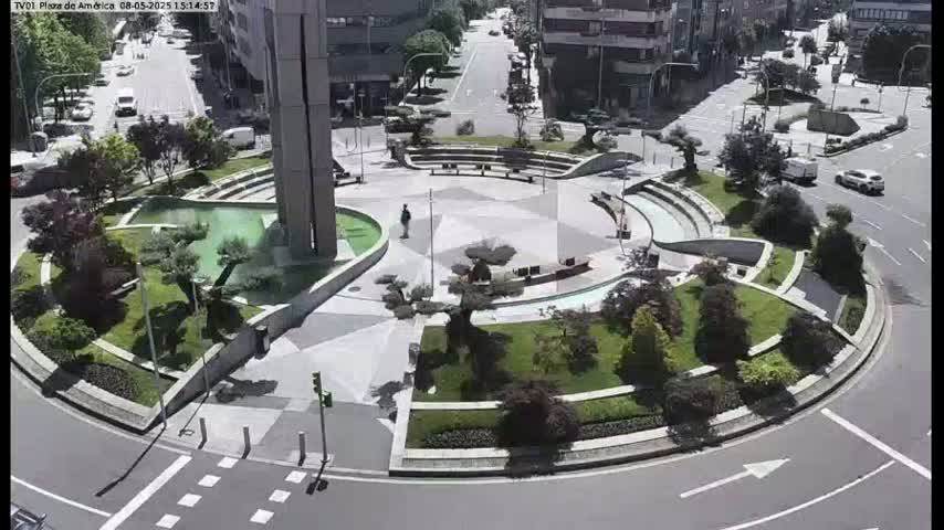 A circular plaza with landscaped gardens, a central water feature, and seating areas is surrounded by a roadway with sparse traffic on a sunny day.