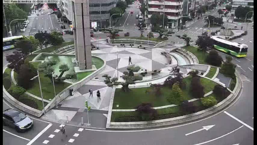 An overcast day reveals a circular plaza with landscaped gardens, geometric pathways, and a few pedestrians, situated near a city street with some cars and a bus.