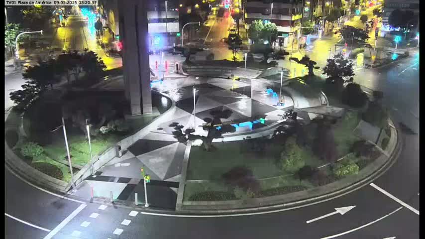 A wet, nighttime view of a plaza with a geometric pattern, surrounded by roadways and landscaping, with minimal vehicle traffic.