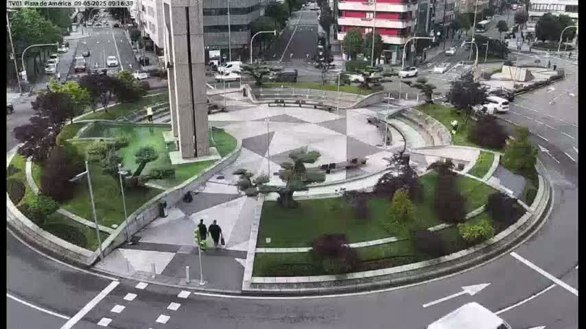A circular plaza with landscaped gardens and geometric paving is surrounded by a roadway with several cars visible on a cloudy day.