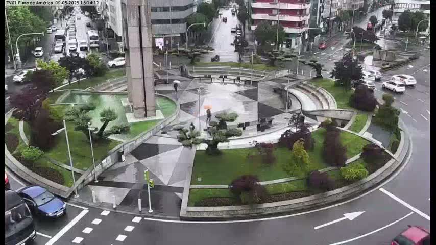 A rain-slicked roundabout, landscaped with geometric patterns and various greenery, is surrounded by a street with cars, and nearby buildings.