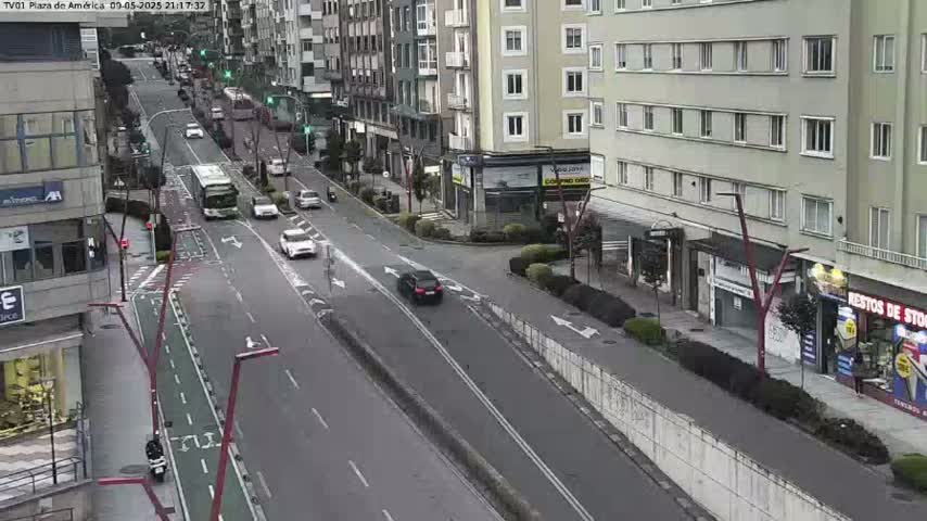 A mostly empty city street scene shows several cars and buses moving along a multi-lane road that curves, passing buildings on either side in overcast daylight.