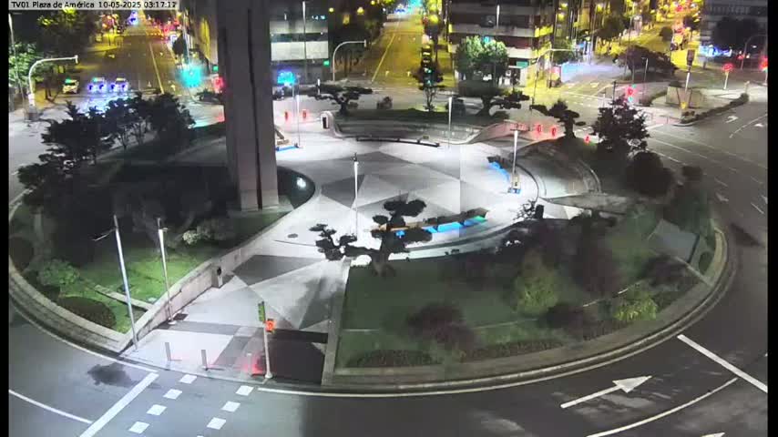 A nighttime view of a paved plaza with landscaping and a roundabout, illuminated by streetlights.