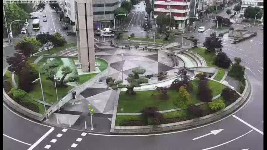 A rain-soaked roundabout features a geometrically designed plaza with landscaping and several small trees, surrounded by city streets and buildings.