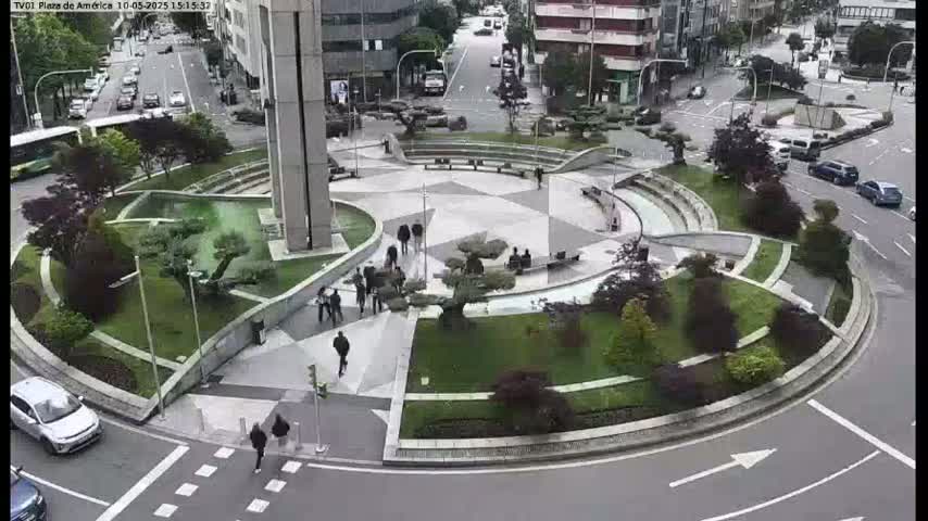 A circular plaza with landscaped gardens and seating areas is surrounded by roadways and buildings, with several people walking around on a sunny day.