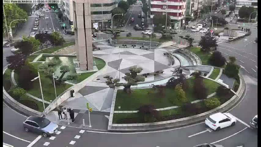 A paved roundabout with landscaping and several cars is visible from an overhead perspective, under a clear sky.
