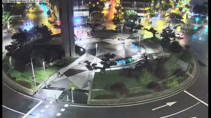 A nighttime aerial view of a landscaped traffic circle with wet pavement, showing several vehicles on surrounding roads.