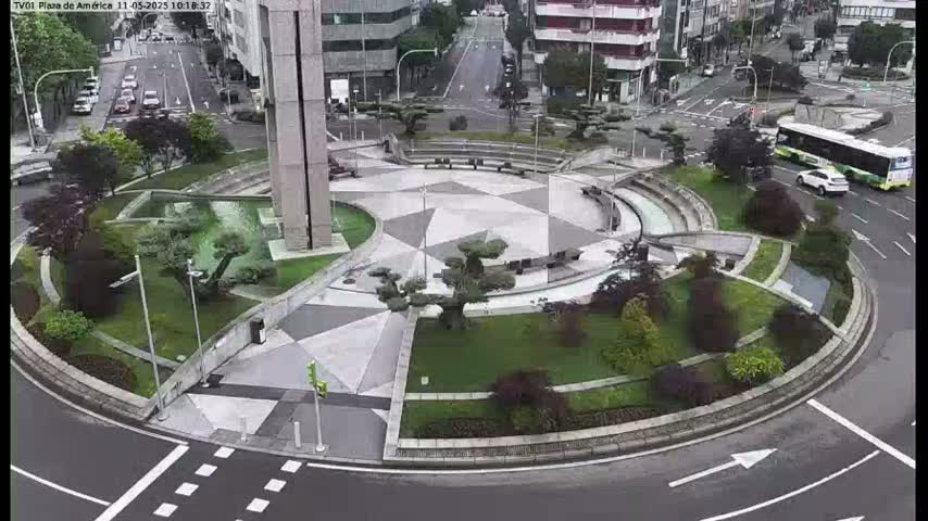 A circular plaza with geometric landscaping and a central fountain is surrounded by roads and buildings, with a bus and several cars visible, under overcast skies.