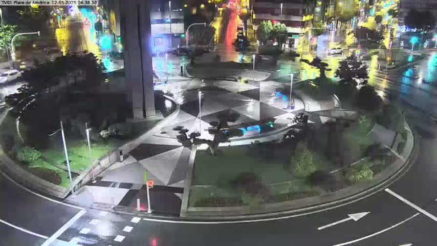 A nighttime aerial view of a plaza with a geometrically patterned paved area, surrounded by landscaping, and wet roads indicating rain.