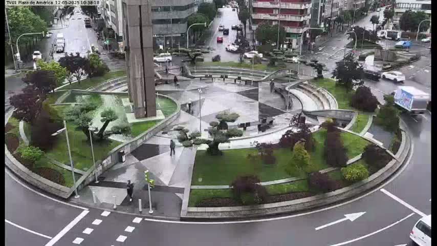 A rain-slicked roundabout features a landscaped center with geometric patterns, small trees, and seating areas, surrounded by a road with cars and pedestrians.
