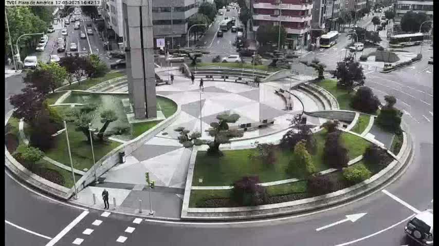 A circular plaza with landscaped gardens and a geometric stone pavement is surrounded by a roadway with cars, under an overcast sky.