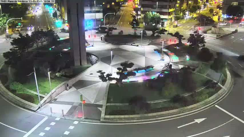 A nighttime aerial view of a landscaped roundabout with geometric paving, illuminated by streetlights, surrounded by city buildings and roads with some cars visible.