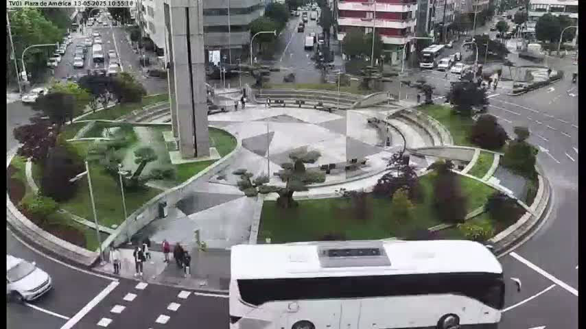 A white bus approaches a circular plaza with landscaped gardens and a central paved area, under overcast skies.