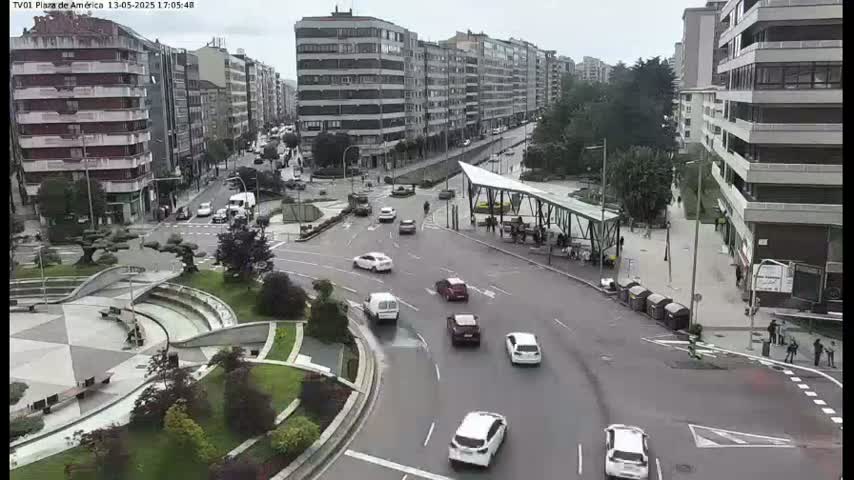 An overcast day shows a plaza with several cars driving through an intersection, surrounded by multi-story buildings.