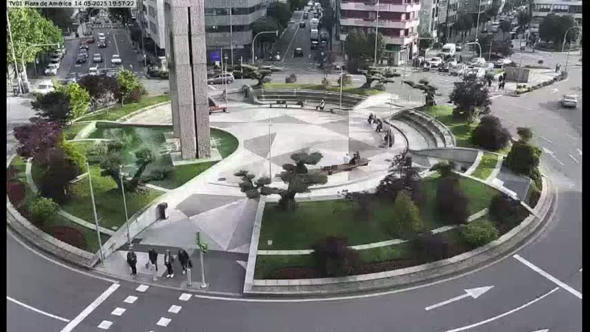 An aerial view shows a landscaped traffic circle with a central plaza and surrounding buildings, on a partly cloudy day.