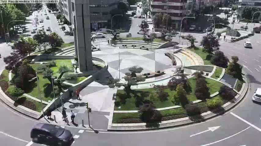 A landscaped traffic circle features a central plaza with benches and greenery, surrounded by a roadway on a sunny day.
