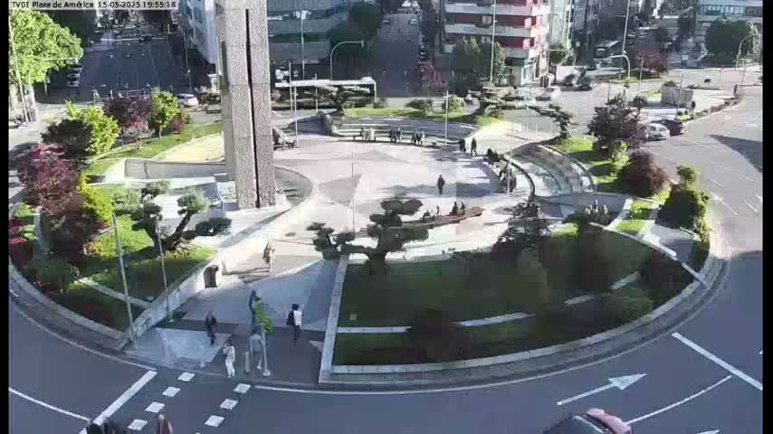 A sunny aerial view of a roundabout in a city, featuring landscaping with various trees and shrubs, pedestrians, and vehicles.