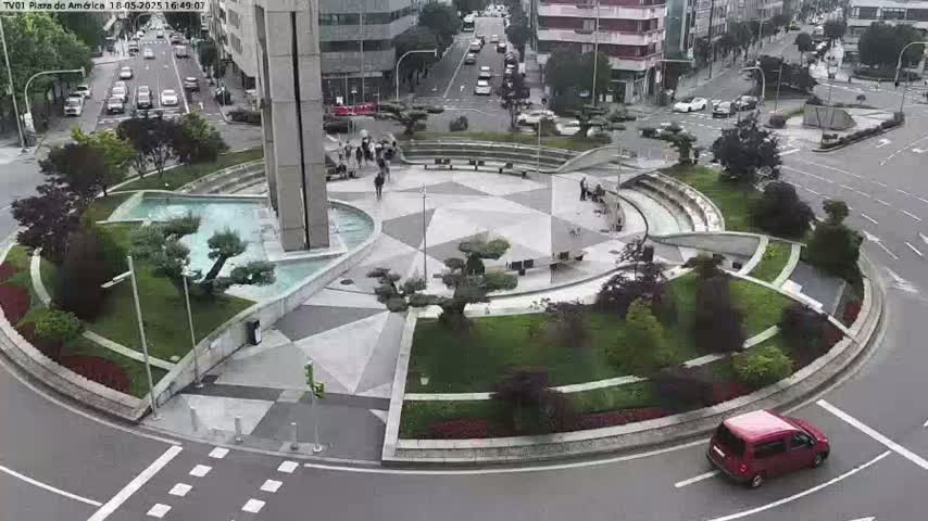 An aerial view of a circular plaza with a fountain, landscaping, and several people walking, situated in the middle of a roadway on an overcast day.