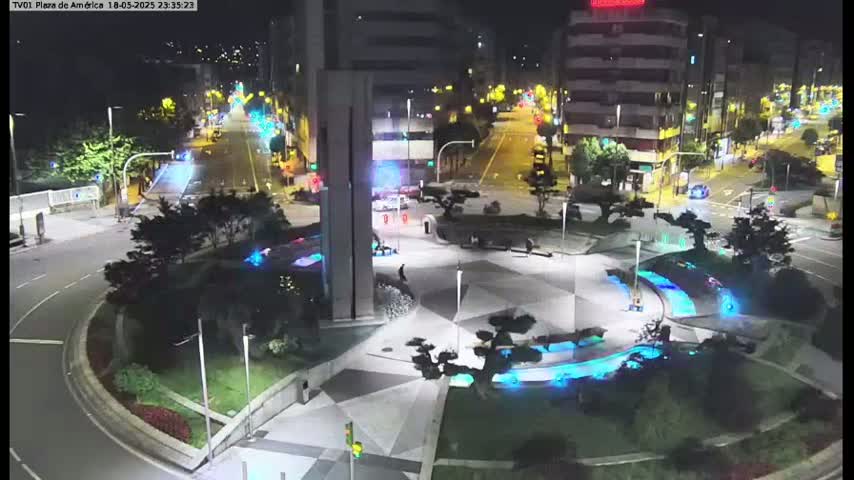 A nighttime aerial view of a plaza with geometric landscaping features illuminated in blue, surrounded by city streets and buildings.