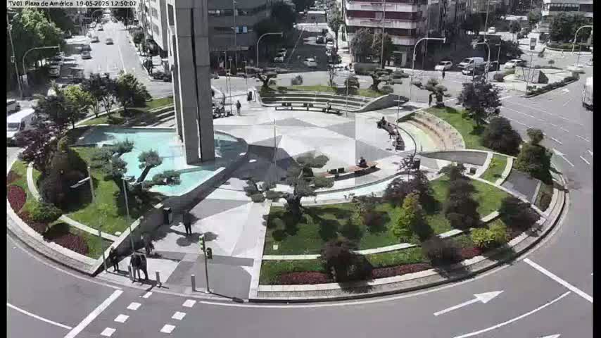 A landscaped roundabout with a central fountain and several people walking around, on a sunny day.