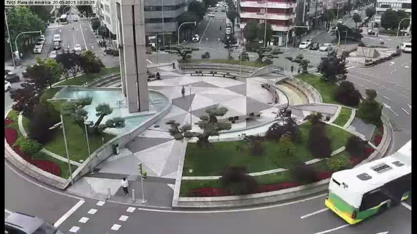 An overhead view of a roundabout with a geometrically patterned plaza at its center, surrounded by landscaping and buildings, on an overcast day.