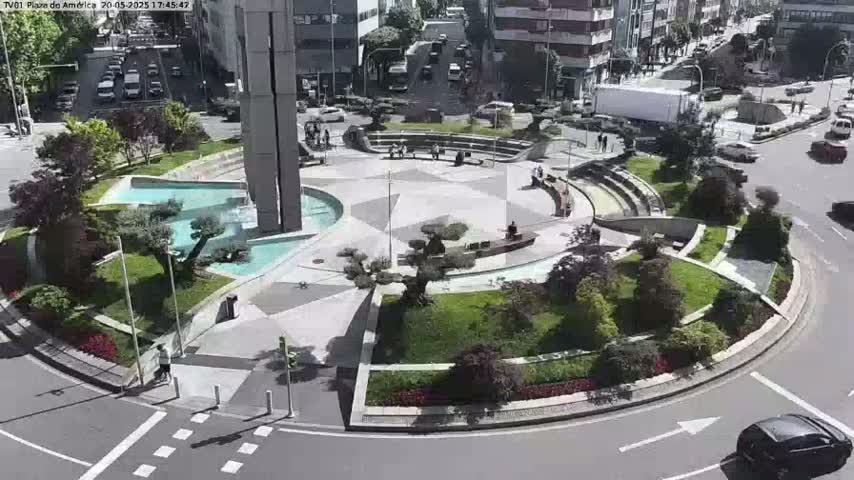 A circular plaza with a fountain, landscaping, and seating areas is situated in a city under sunny skies, with cars visible on surrounding roads.
