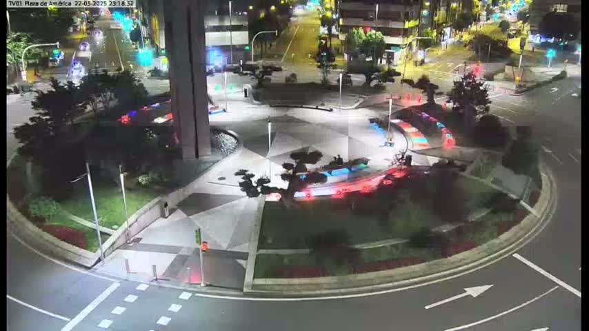 A nighttime aerial view of a roundabout with a geometrically patterned center, surrounded by landscaping and streetlights, with cars visible on the roads.