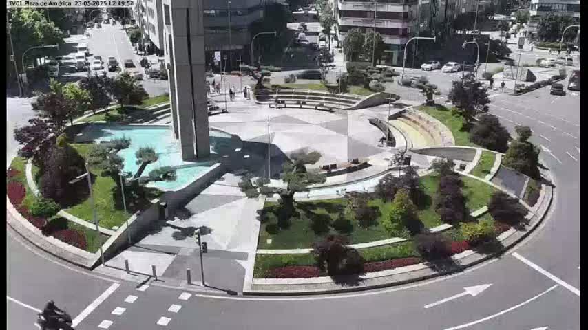 A circular plaza with landscaping features, including fountains and trees, is surrounded by a roadway under sunny conditions.